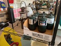 Box containing six green Bernzomatic propane canisters with white labels on a metal shelf, alongside a metal propane lantern with a rusted finish and capped wick inside.