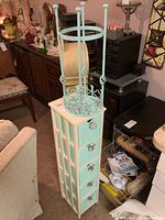 Photo showing tall narrow pastel turquoise storage drawer tower with five drawers and decorative knobs, topped with a turquoise metal stool or plant stand with florally cut-out design