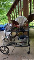Full view of metal two-tier teacart loaded with various items, showcasing mesh shelves, larger rear wheels, smaller front wheels, outdoors setting.