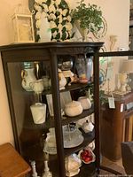 Display cabinet with white ceramic teapot including gold handles, matching creamer jug, multiple covered dishes, clear tiered dessert server, and small decorative items such as white candle holders.