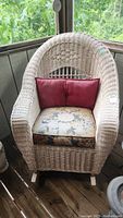 Full view of wicker rocking chair with red pillow and floral seat cushion. Shows weathered wicker.