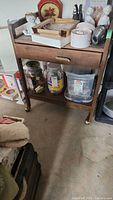Full view of the wooden cart with drawer, top surface containing various containers and decorative items, and bottom shelf with clear plastic containers and jars.
