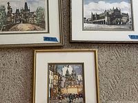 Three framed European city art pieces displayed on carpet, showing detailed old city architecture and street scenes