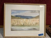 Framed watercolour painting showing a cliff with water and hills under a cloudy sky. The frame is gold with white matting. Price tag visible showing $75.