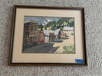 Full view of the framed watercolour painting depicting rustic wooden buildings in a village setting with a forest background. Shows overall condition and framing.