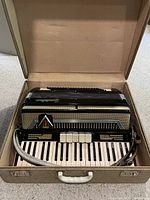 Black and cream Hohner piano accordion inside a beige carrying case, showing the instrument's keyboard, bass buttons, and strap.