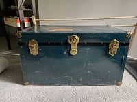 Front view of the teal metal trunk showing brass latches and corner protectors with visible scratches and wear on surface.