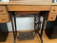 Front view of vintage Singer sewing machine built into wooden sewing cabinet with drawers and treadle base showing wear.