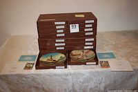 View of 16 brown boxes stacked with labels naming baseball legends, with two collector plates displayed in front, plates painted with baseball player portraits and certificates of authenticity alongside.