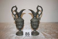 Photo showing the matching pair of antique metal Grecian ewer pitchers with dragon handles and face designs on the belly.