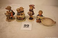 Photo of all four Hummel figurines arranged in a row on a white cloth background with lot number 99 card visible