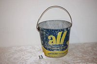 Front view of blue and yellow galvanized metal bucket with ALL detergent logo, placed on a patterned surface.