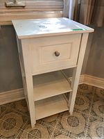 Front view of white wooden cabinet showing single drawer and two shelves below, surface wear visible