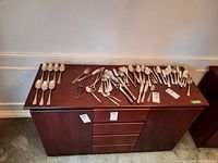 Full view of all cutlery items displayed on wooden cabinet including spoons, forks, knives and serving tongs.