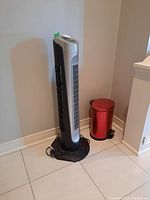 Photo showing Holmes tower fan standing on black base beside a small red metal pedal trash can against corner wall on tiled floor.