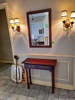 Wood hall table with two drawers, one handle missing, placed beneath a framed wall mirror with decorative corner accents. Two wall sconces flank the mirror.