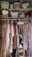 Closet with assorted clothing including tops, shirts in dry-cleaning plastic covers and hanging loose in various colors; shelf above contains storage baskets and sewing box.