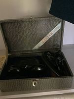 Opened textured case containing a black ionizer device with two bulbous ends, coiled wiring on right side, blue velvet interior lining, and a label reading 'IONIZER' inside lid.