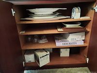 Wooden cabinet shelves holding various ceramic plates stacked including fish-shaped and rectangular styles, a few clear glasses, and decorative boxes at bottom shelf.