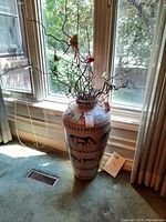 Full view of tall decorative urn with faux branches and birds near window, placed on carpeted floor.