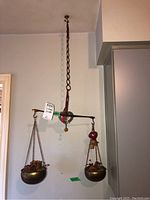 Full view of vintage hanging metal balance scale mounted from ceiling, showing the chain, arm, and two brass weighing pans holding decorative items.