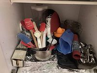 Cupboard shelf with assorted baking tools including spatulas, measuring spoons, and kitchen utensils grouped in bowls and containers with other items stacked behind.