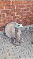 Two-piece concrete birdbath with base and basin leaning against a brick wall on paved surface