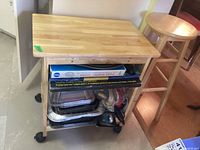 View of wood cart on castors showing rectangular surface, single shelf below with assorted items stored, and black castors on each leg.