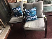 Two patio chairs with tan cushions and blue patterned pillows side by side on porch.