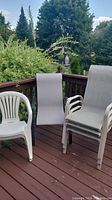 Three garden chairs on a wooden deck: one white plastic chair and two grey fabric sling chairs with metal frames, one stack of sling chairs visible.