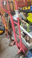 Side view of red steel hand cart showing the curved handle and pneumatic tire on concrete floor next to woodworking tools.