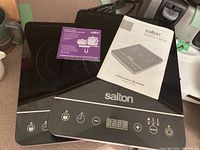 Two black Salton induction cooktops on countertop. Instruction booklet placed on top of one unit. Both feature LCD displays and touch control buttons.