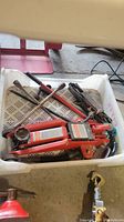 Plastic crate containing a red hydraulic floor jack, a black mechanical screw jack, and a rusty four-way wheel lug wrench.