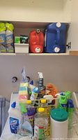 Shelf with blue and red liquid detergent jugs, Lysol sanitizer, cleaning sponges, spray bottles, and assorted bottles in front of washing machine.