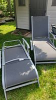 Two patio loungers with gray mesh fabric and silver metal frames placed on grass in front of a white outdoor building.