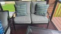 Front view of outdoor patio loveseat showing gray cushions and striped blue-green pillows, metal frame is visible with armrests