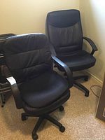 Two black faux leather office chairs side by side in a small room on carpet.