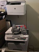 Brother DCP 7040 printer stacked below HP LaserJet M180 printer wall-mounted on beige wall, two gray desktop computer speakers on top of Brother printer, several boxes nearby.