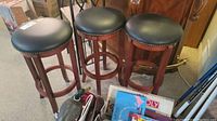 Three wooden bar stools with black padded seats and nailhead trim, shown from front and side angles together in a room with various items around.
