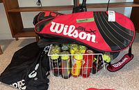 Photo showing the Wilson red and black racket case on the floor alongside wire basket filled with multiple cans of tennis balls and black duffle bags.