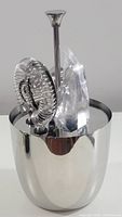 Front view of the silver-tone metal ice bucket with some of the bar tools inside, showing the polished finish and spring coil cocktail strainer.