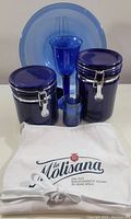 Photo showing 2 blue glass canisters with metal clasps, blue glass goblet and votive holder, light blue glass plate, and white La Molisana apron