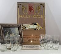 Photo of the entire lot showing six decorated glasses, two framed prints, postcards laid out, and a large vintage Rolls-Royce advertising sign