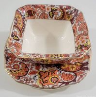 Squared bowl stacked on matching underplate showing paisley border in warm colors with gold trim