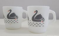 Front and side view of two vintage white milk glass mugs featuring grey swan pattern with orange accents, showing size and design details.