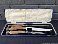 Close-up view of three-piece meat carving set in fitted case showing stag horn handles and silver ornate ends, carving knife with curved blade, fork, and sharpening steel for meat carving.