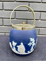 Front view showing blue Jasperware with white classical relief figures and foliage, EPNS metal lid and handle, wooden knob on top.