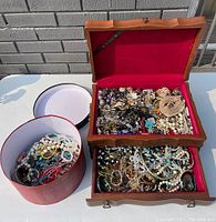 Open two-tier wooden jewelry box showing red velvet-lined compartments filled with assorted jewelry and round hat box beside it
