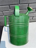 Front view of green galvanized steel watering can against gray brick wall, showing cylindrical body, top carry handle, side pour handle, and brass spout.
