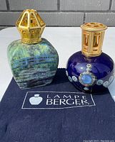 Two Lampe Berger fragrance lamps positioned on a black cloth with Lampe Berger branding, shown outdoors in natural light.